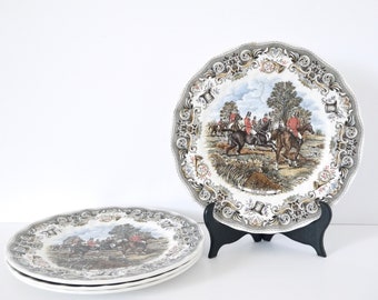 May include: Three white ceramic dinner plates with a brown and green floral border. The center of the plates feature a hunting scene with riders on horseback. The text "The First" is printed on the bottom of the plate.