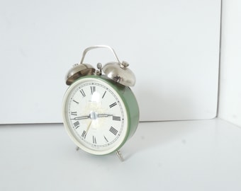 Puede incluir: Un reloj despertador vintage verde con campanas plateadas y números romanos en la esfera.