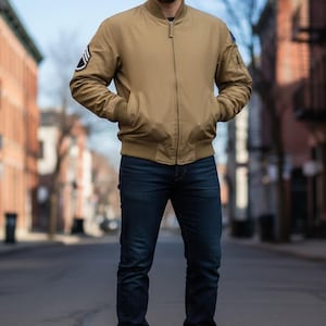 May include: A tan bomber jacket with a zip fastening, paired with dark blue jeans and brown boots. The jacket features a ribbed collar and cuffs, and a patch on the left sleeve. The person is standing on a city street.