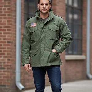 May include: An olive green military-style jacket with an American flag patch on the chest. The jacket features multiple pockets, a high collar, and the letters "U.S." printed on the right side. The person is wearing blue jeans.