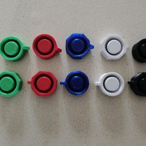 Op de afbeelding: Een verzameling van tien kleine, ronde objecten in verschillende kleuren: groen, rood, blauw, wit en zwart. Elk object heeft een cirkelvormig midden en een klein handvat.