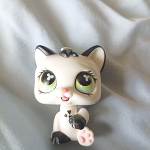 Peut inclure: Figurine de chat blanc avec des détails noirs sur les oreilles, les pattes et la queue. Le chat a de grands yeux expressifs avec des détails verts et bleus. La figurine a un nez et une bouche roses, et un petit nœud noir sur la tête.