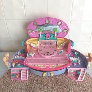 Può includere: Un playset vintage a forma di casa delle bambole nei toni del rosa, viola e giallo. Il playset presenta più stanze, scale ed elementi decorativi, tra cui uno specchio e dettagli a forma di stella. La casa delle bambole è progettata per aprirsi e chiudersi.