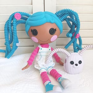 Peut inclure: Une poupée aux cheveux bleus en spirale, yeux en boutons noirs et joues roses, portant une robe salopette blanche sur un haut rose à manches longues et des baskets bleues. À côté se trouve un petit sac rond blanc avec des yeux en boutons noirs.