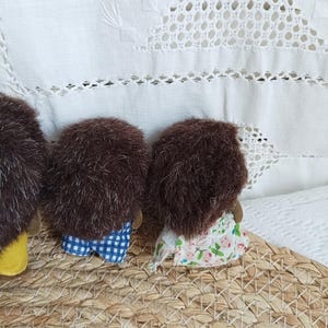 May include: Three handmade hedgehog dolls with brown fur and fabric clothing. One doll wears blue and white checkered shorts, another a floral dress. The dolls are arranged on a woven basket, with a white, embroidered background.