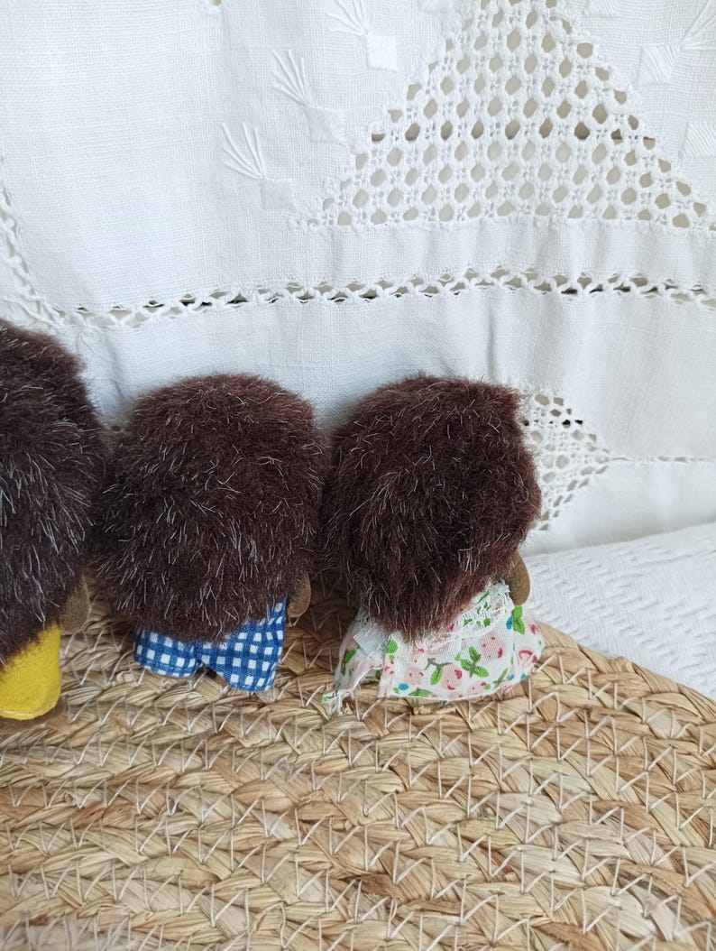 May include: Three handmade hedgehog dolls with brown fur and fabric clothing. One doll wears blue and white checkered shorts, another a floral dress. The dolls are arranged on a woven basket, with a white, embroidered background.