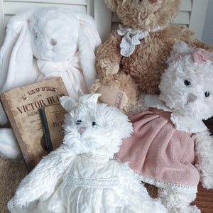 Puede incluir: Una colección de animales de peluche, incluyendo un conejo blanco con largas orejas, un oso de peluche marrón y un oso blanco con un vestido rosa. Un libro vintage con texto en francés es visible. Los juguetes son suaves y esponjosos.