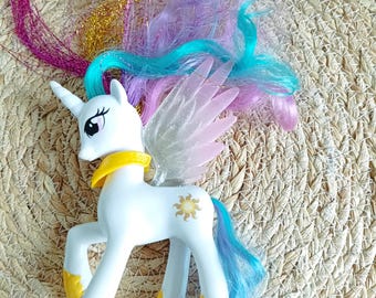 May include: A white toy pony with a horn, wings, and colorful hair. The pony has a yellow collar, hooves, and accents. The mane and tail are a mix of blue, purple, and pink. The wings are translucent pink.