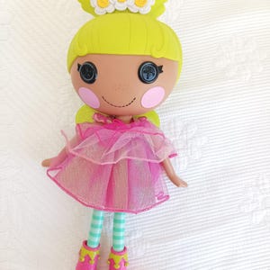 Peut inclure: Une poupée aux cheveux jaune vif, une couronne de fleurs vert citron et des yeux en forme de boutons noirs. Elle porte une robe en tulle rose à plusieurs épaisseurs, des leggings rayés vert clair et blanc, et des bottes roses à bords festonnés.