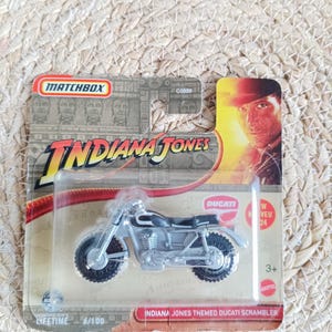 Peut inclure: Moto jouet Ducati Scrambler sur le thème d'Indiana Jones de Matchbox, dans un emballage plastique transparent. La moto est argentée et noire. L'emballage présente le logo et l'image d'Indiana Jones.