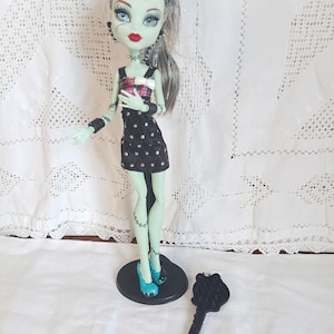 Peut inclure: Poupée à la peau verte, aux cheveux gris et noirs et au rouge à lèvres rouge. La poupée porte une robe noire à pois blancs, un haut rose et noir et des chaussures turquoise. Une brosse noire est sur la surface blanche.