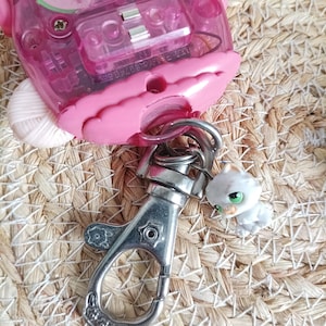 May include: A pink electronic toy with a clear plastic window, a silver metal clasp, and a small white cat figurine with green eyes. The toy is on a woven straw surface.
