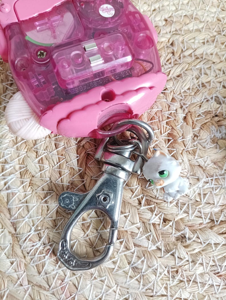 May include: A pink electronic toy with a clear plastic window, a silver metal clasp, and a small white cat figurine with green eyes. The toy is on a woven straw surface.