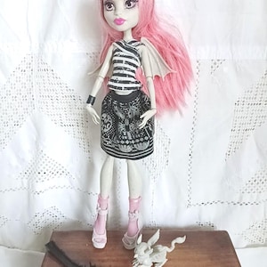 Peut inclure: Une poupée Monster High avec des cheveux roses, un haut rayé noir et blanc et une jupe noire avec un motif de crâne. La poupée a la peau pâle, du rouge à lèvres rose et porte des chaussures roses. Accessoires : une petite créature blanche et une brosse noire.