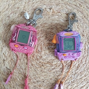 May include: Two vintage LPS electronic pet keychains. One is pink with a heart on the screen, the other is purple and blue with a tree graphic. Both have silver keyrings and small accessories.