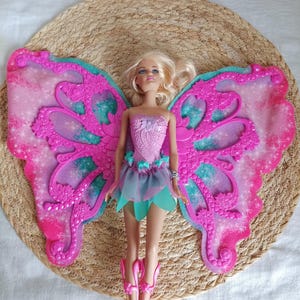 Op de afbeelding: Een pop met blond haar, gekleed in een roze lijfje, een blauw en groene rok en roze laarzen. De pop heeft grote roze en blauwe vlindervleugels. De pop ligt op een geweven, ronde mat.