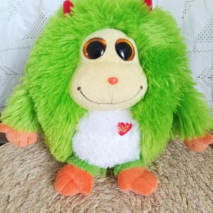 Peut inclure: Une créature en peluche ronde avec une fourrure verte ébouriffée, de grands yeux marron et des mains et pieds orange. Elle présente un visage crème, un ventre blanc, deux petites cornes rouges et un écusson en forme de cœur avec LUV ME.