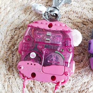 May include: A pink, translucent toy with a silver metal clip. The toy has a house-like shape with a pink roof and a clear body revealing internal components. The text "&copy; 2005 Hasbro MADE IN CHINA" is visible. A pink string is attached.