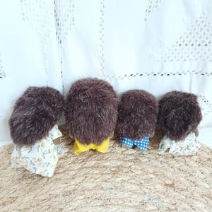 May include: Four small, brown, plush hedgehog figurines. Each has a different outfit: a floral dress, yellow shorts, blue checkered shorts, and a floral dress. They are arranged on a woven mat.