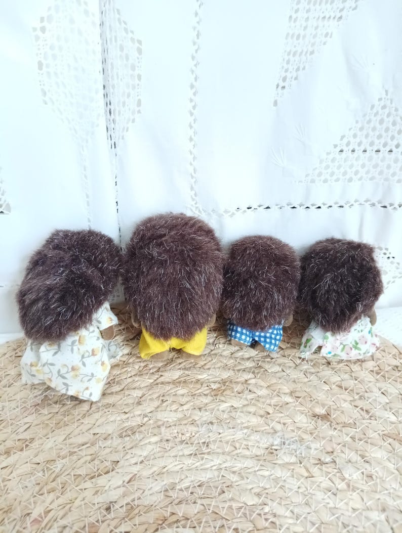 May include: Four small, brown, plush hedgehog figurines. Each has a different outfit: a floral dress, yellow shorts, blue checkered shorts, and a floral dress. They are arranged on a woven mat.