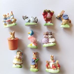 Pode incluir: Uma coleção de figuras de cerâmica com vários personagens animais. As figuras incluem coelhos, um pato e um gato, todos em trajes coloridos e detalhados. Cada figura é colocada em uma pequena base gramada, criando uma exibição caprichosa.