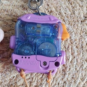 May include: A vintage handheld electronic toy with a clear blue center and a purple frame. The toy has an orange accent and a metal clip. The text "U.S. Pat. No. 6971855" and "&copy; 2006 Hasbro, MADE IN CHINA" are visible.