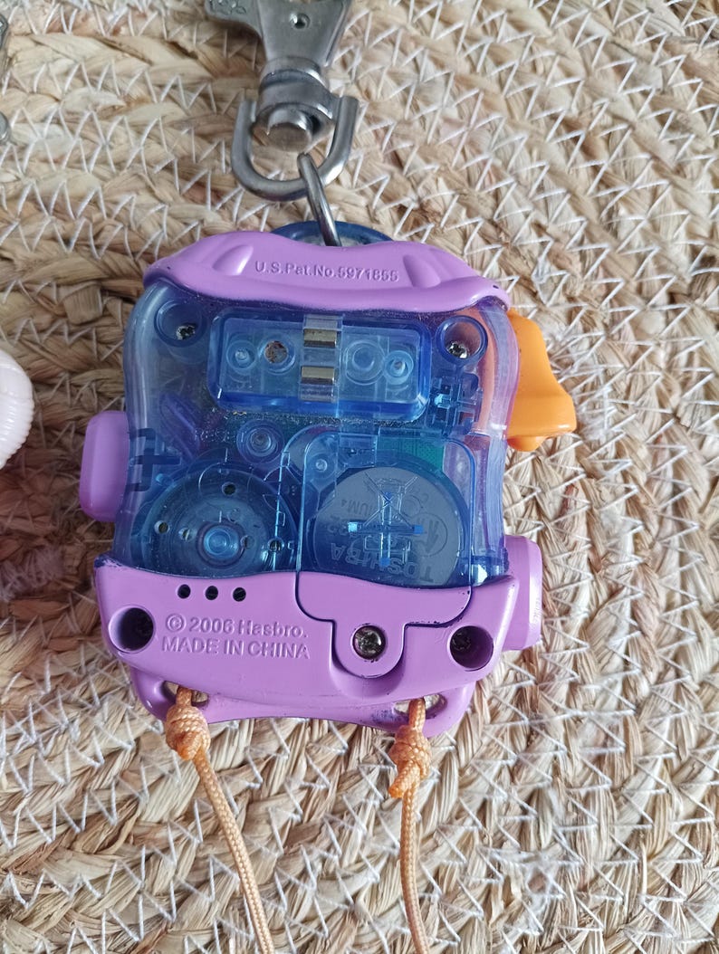 May include: A vintage handheld electronic toy with a clear blue center and a purple frame. The toy has an orange accent and a metal clip. The text "U.S. Pat. No. 6971855" and "&copy; 2006 Hasbro, MADE IN CHINA" are visible.