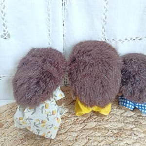 May include: Three handmade hedgehog dolls with brown fur and fabric clothing. One doll wears a floral dress, another a yellow outfit, and the third a blue and white checkered pair of shorts. The dolls are arranged on a woven surface.