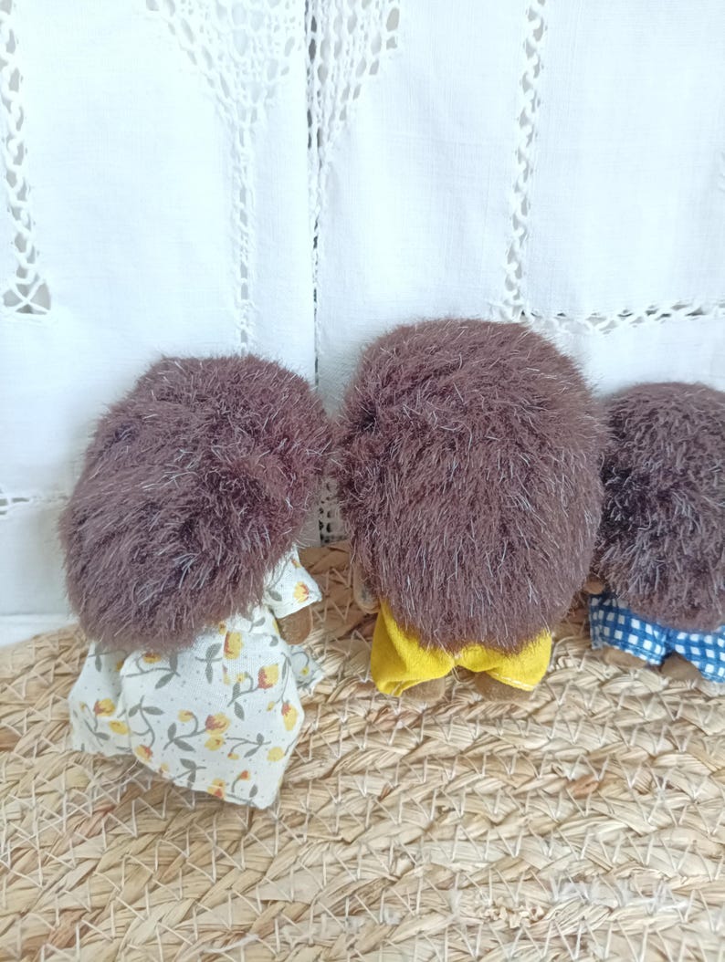 May include: Three handmade hedgehog dolls with brown fur and fabric clothing. One doll wears a floral dress, another a yellow outfit, and the third a blue and white checkered pair of shorts. The dolls are arranged on a woven surface.