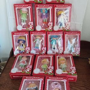 Może przedstawiać: Kolekcja lalek Strawberry Shortcake w oryginalnych pudełkach. Każda lalka znajduje się w czerwonym pudełku z białym, koronkowym obramowaniem i napisem "Strawberry Shortcake" oraz "The Original 1980's Design". Lalki mają kolorowe stroje.