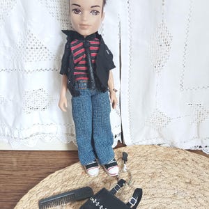 Könnte beinhalten: Bratz-Puppe mit schwarzer Jacke über einem rot-schwarz gestreiften Hemd, blauen Jeans und Turnschuhen. Zubehör: schwarze Handtasche, Kamm und silberne Kette. Die Puppe steht auf einer gewebten Matte.