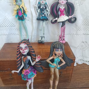 Peut inclure: Cinq poupées Monster High sont présentées sur une boîte en bois. Les poupées ont des designs uniques, notamment des tenues, des coiffures et du maquillage colorés. Les poupées sont dans diverses poses, mettant en valeur leurs styles et personnalités individuels. Les poupées sont un objet de collection populaire.