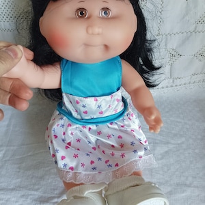 Peut inclure: Une poupée vintage aux cheveux noirs, aux joues roses et aux yeux bicolores. La poupée porte un haut bleu et une robe blanche ornée de cœurs roses et de fleurs bleues. Des chaussures et des chaussettes blanches complètent la tenue. Le bras de la poupée est tenu par une main.