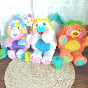 Könnte beinhalten: Drei bunte Plüschtiere. Das erste ist ein Bär mit Regenbogenfell. Das zweite ist ein weißes Häschen mit gelben Haaren und blauen Ohren. Das dritte ist ein orangefarbener Bär mit grünen Haaren. Sie sitzen auf einer runden gewebten Matte.