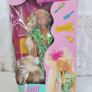 Puede incluir: Una muñeca en una caja con la etiqueta "HAIR STYLE" con el texto "Steffi Love". La muñeca tiene pelo largo rubio, ojos azules y viste un conjunto de lunares verdes y blancos. La caja incluye accesorios para peinar el cabello.