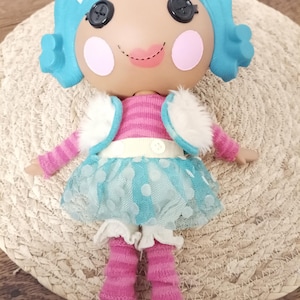 May include: A doll with bright blue hair, black button eyes, and pink circular cheeks. The doll wears a pink and white striped long-sleeve shirt, a white faux fur vest, a blue polka-dot tulle skirt, and pink leg warmers over white boots.