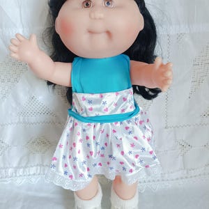 Puede incluir: Una muñeca vintage con cabello negro, ojos marrones y mejillas rosadas. La muñeca lleva una blusa turquesa y un vestido blanco adornado con corazones y flores rosas y azules. El vestido tiene un ribete de encaje blanco. La muñeca lleva calcetines blancos y zapatos blancos.