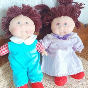 Peut inclure: Deux poupées vintage avec des cheveux en fil et des visages peints. L'une porte une combinaison bleue avec un col blanc et des chaussures rouges. L'autre est vêtue d'une robe blanche avec un nœud violet et des chaussures rouges. Les poupées sont sur un tapis tissé.