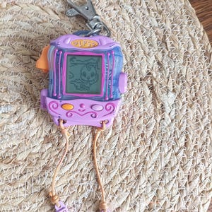 May include: A vintage purple and blue handheld electronic pet game with a silver keychain clip. The game features a small screen displaying a pixelated animal. The game has the text "LPS" on the top. Attached are two light brown cords with small purple accessories.