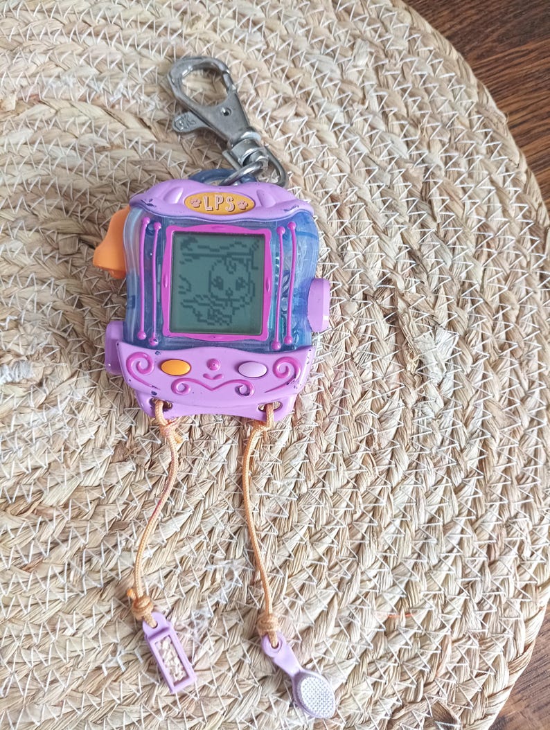 May include: A vintage purple and blue handheld electronic pet game with a silver keychain clip. The game features a small screen displaying a pixelated animal. The game has the text "LPS" on the top. Attached are two light brown cords with small purple accessories.