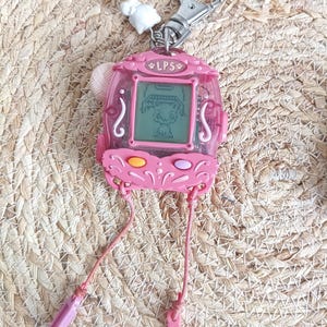 May include: Pink LPS handheld electronic game with a digital screen displaying a cartoon character. The game has a silver keychain clip and pink straps with plastic accessories. The text "LPS" is visible on the top of the device.