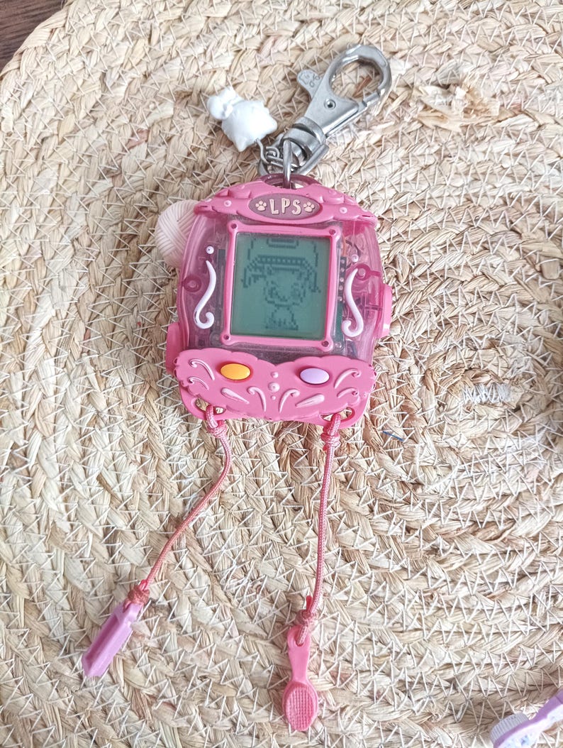 May include: Pink LPS handheld electronic game with a digital screen displaying a cartoon character. The game has a silver keychain clip and pink straps with plastic accessories. The text "LPS" is visible on the top of the device.