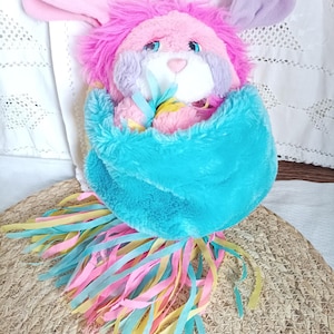 Peut inclure: Une peluche en forme de lapin avec une fourrure rose, violette et bleue, enveloppée dans un tissu bleu turquoise. Le lapin a de longues oreilles et des yeux bleus. Des rubans colorés roses, jaunes et bleus pendent du bas.