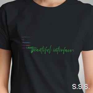 Op de afbeelding: Zwart T-shirt met groene en paarse tekst. Het shirt heeft codefragmenten en de zin "beautiful interface" in een cursief lettertype. De rechter benedenhoek heeft de letters "S.S.S." in een pixelachtige stijl.