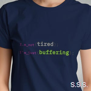 Pode incluir: Camiseta azul marinho com o texto "I'm not (tired); I'm just (buffering);" em uma fonte de estilo de código. O texto é em rosa e verde. A camiseta também tem as letras "S.S.S." no canto inferior direito.