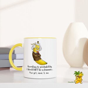 Può includere: Tazza in ceramica bianca con manico e bordo gialli. La tazza presenta un cartone animato di Albert Einstein come una banana con il testo "Secondo la probabilità, NON dovrei essere una banana... Eppure, eccomi qui."