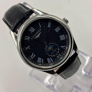 Könnte beinhalten: Eine Longines Automatikuhr mit rundem, silberfarbenem Edelstahlgehäuse und strukturiertem, dunkelblauem Zifferblatt. Das Zifferblatt verfügt über silberne römische Ziffern als Stundenmarkierungen, eine Mondphasenanzeige und ein schwarzes Lederarmband.