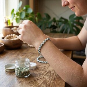Könnte beinhalten: Ein Perlenarmband mit kleinen, runden, grün-weißen Perlen in einem gedrehten Muster. Das Armband wird am Handgelenk getragen, und ein durchsichtiges Glasgefäß mit passenden losen Perlen steht in der Nähe auf einer hölzernen Oberfläche.