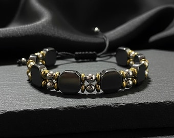May include: A black beaded bracelet with square and round beads. The bracelet features dark gray square beads, interspersed with gold and silver-colored round beads. The bracelet is on a black surface.