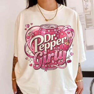 May include: Cream-colored t-shirt featuring a pink Dr. Pepper can design with the words "Dr. Pepper Girly" in a playful font. The design includes hearts, cherries, and ribbons, creating a fun and feminine aesthetic.